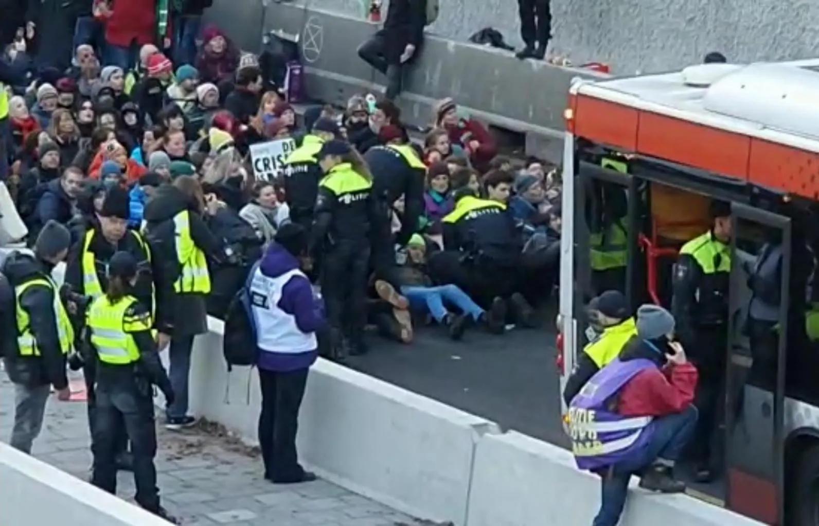 The picture is taken from a high angle. To the left is a crowd of people sitting on the ground in a tight group. Standing in front of them are about 5 cops. Two are trying to pull an activist out of the group. In the foreground to the right is the back of a bus with police officers and a photographer.