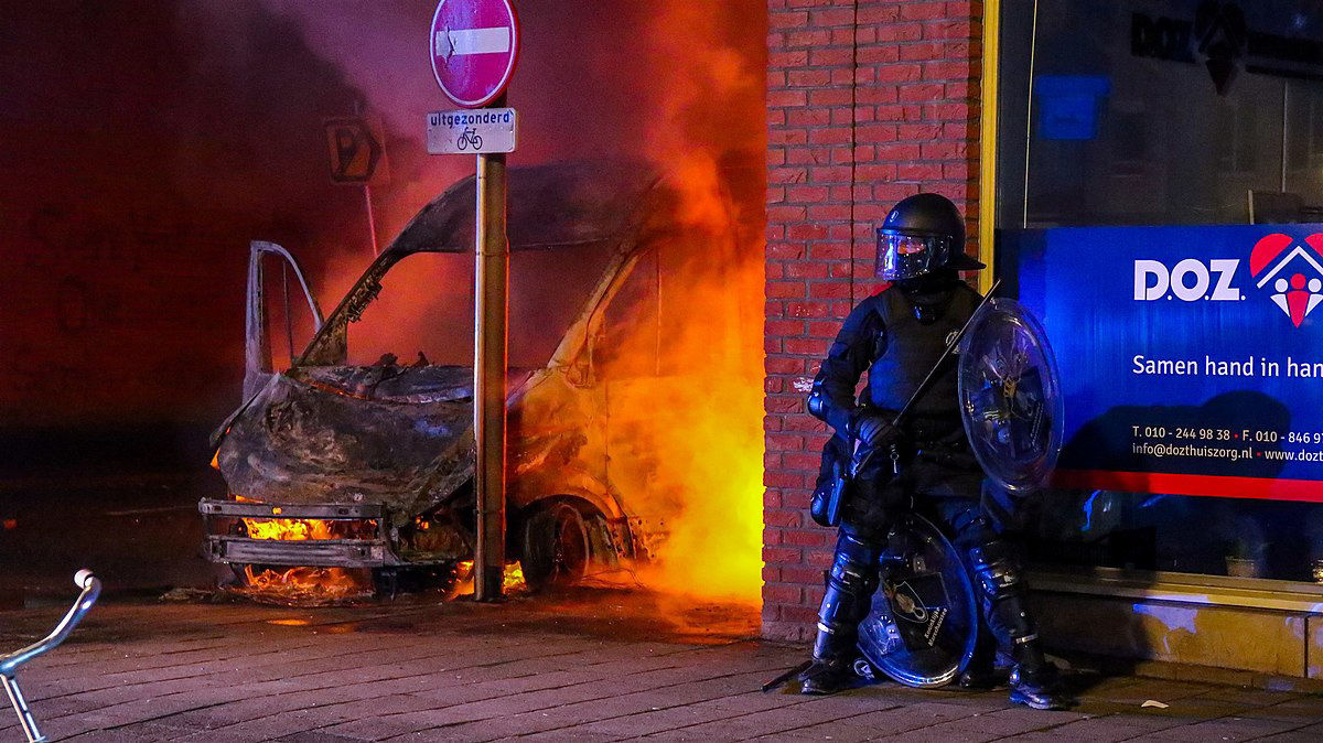 A police officer wearing heavy armour on a street corner, with around the corner a burnt out van with visible flames next to it.
