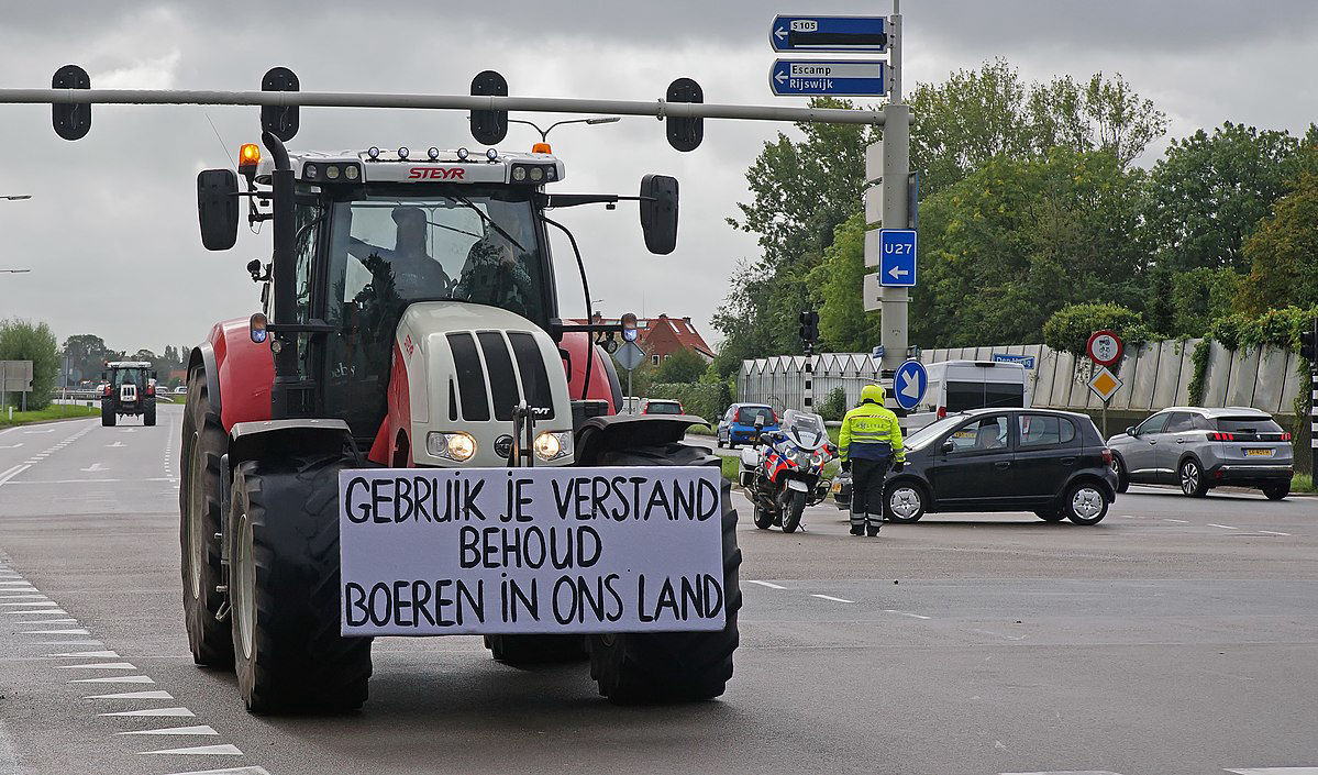 A tractor on a crossroad, with a sign tied to the front that reads 'Use your brain, keep farmers in our country' in Dutch.